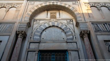 Decorated entrance with gray-and-white striped arches, marble columns, carved capitals, and an Arabic inscription panel.