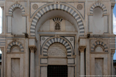 Symmetrical stone façade with a large striped arch, smaller central arch, inscription panel, paired columns, and side niches.