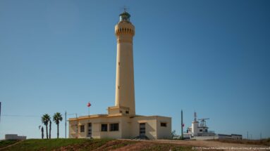 El Hank Lighthouse with tall tapering tower, lantern gallery, low white base buildings, palms, and a Moroccan flag.