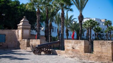 Historic cannon beside low stone walls and palms in a landscaped fortified enclosure with Moroccan flags.