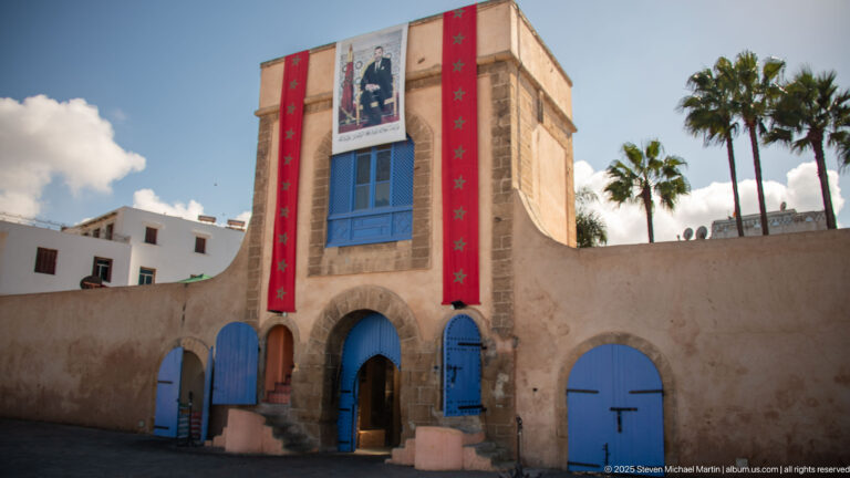 Stone gateway with blue arched doors, red Moroccan banners, royal portrait, and palm trees behind.