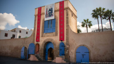 Stone gateway with blue arched doors, red Moroccan banners, royal portrait, and palm trees behind.