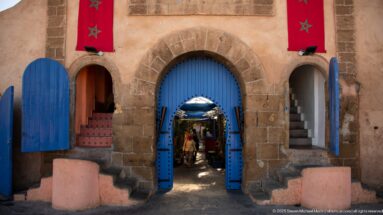 Three stone arches with bright blue doors and Moroccan flags, the central doorway opening into a shaded passage.