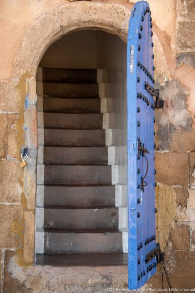 Open studded blue door beneath a stone arch reveals a steep interior stairway.
