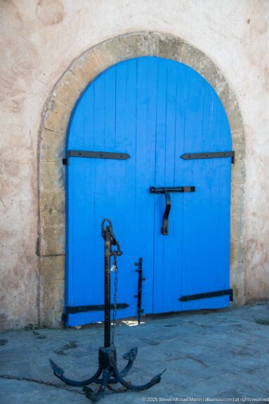 Large iron anchor stands before closed bright-blue double doors set in a shallow stone arch.