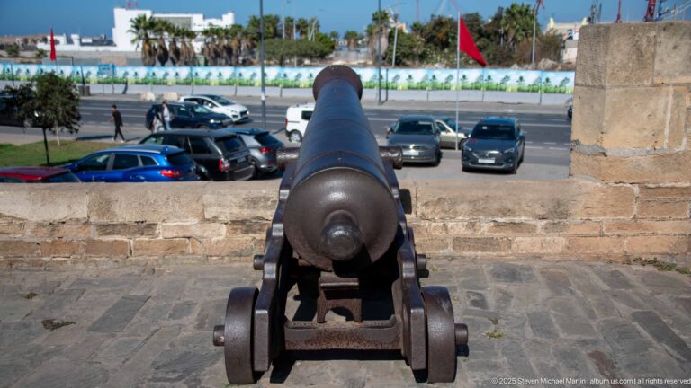 Cannon points over a low stone wall toward parked cars, pedestrians, palms, and a Moroccan flag.