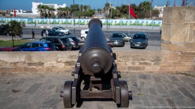 Cannon points over a low stone wall toward parked cars, pedestrians, palms, and a Moroccan flag.