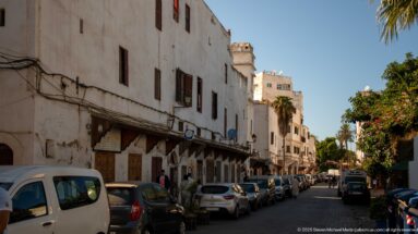 Narrow street lined with weathered white buildings, shopfronts, parked cars, and pedestrians.