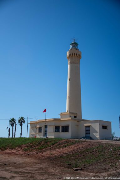 El Hank Lighthouse rising from a long low white building on open ground, with palms and a small Moroccan flag.