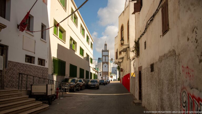 Narrow white-walled lane with green shutters and parked vehicles framing a distant minaret.