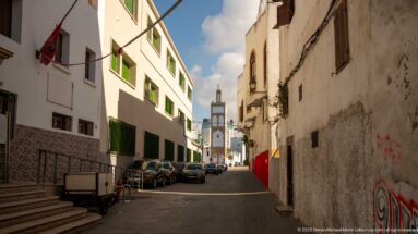 Narrow white-walled lane with green shutters and parked vehicles framing a distant minaret.