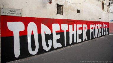 Large white words “TOGETHER FOREVER” painted across a long red-and-black wall beside a sidewalk.