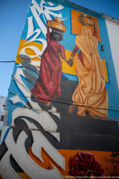 Colorful wall mural of two seated women in red and ochre garments carrying vessels on their heads.