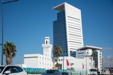 White high-rise tower rises behind a lower civic complex with Moroccan flags, palms, and a slender square tower.