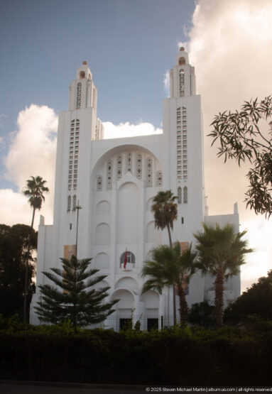 White former Sacred Heart Church with paired towers, broad central arches, palms, and landscaped grounds.