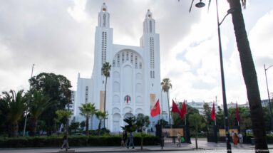 Former Sacred Heart Church framed by palms, trees, Moroccan flags, and pedestrians near the entrance.