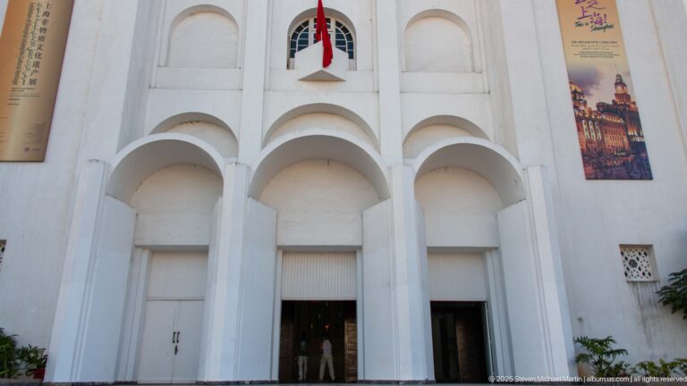 Close view of the former Sacred Heart Church façade with three deep arches, white piers, a Moroccan flag, and exhibition banners.