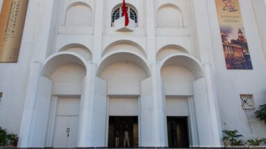 Close view of the former Sacred Heart Church façade with three deep arches, white piers, a Moroccan flag, and exhibition banners.
