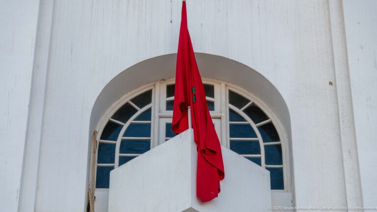 Red Moroccan flag hangs below a large semicircular window in a smooth white building façade.