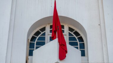 Red Moroccan flag hangs below a large semicircular window in a smooth white building façade.