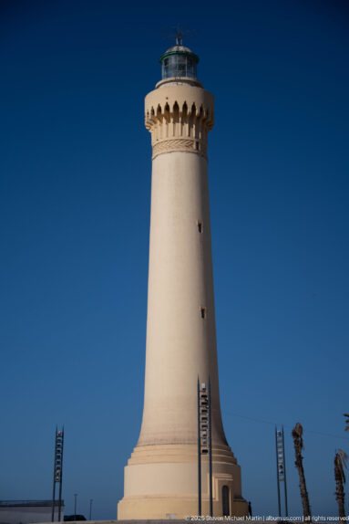 White tapered lighthouse tower with projecting lantern gallery rising against a deep blue sky.