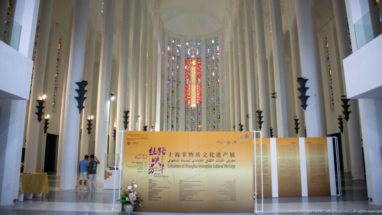 Long white nave of the former Sacred Heart Church leading to tall multicolored stained-glass windows and an exhibition display.