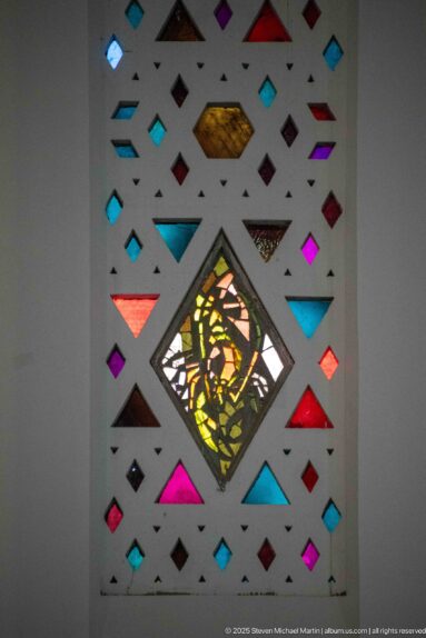 Diamond-shaped figurative stained-glass medallion surrounded by small colorful geometric panes in a pale screen.