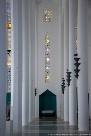 White side aisle with tall piers, narrow colored stained-glass windows, dark wall fixtures, and a low green structure.