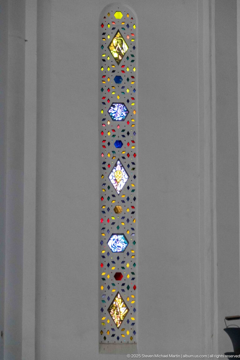Single tall narrow stained-glass window with repeated colored diamond motifs set in a broad white wall.