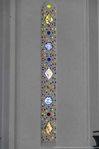 Single tall narrow stained-glass window with repeated colored diamond motifs set in a broad white wall.