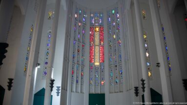 Upward interior view of tall white ribs framing a large multicolored stained-glass wall in the former Sacred Heart Church.