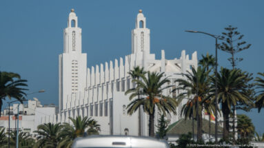 White mosque with twin square towers and vertical façade fins behind palm trees in Casablanca.