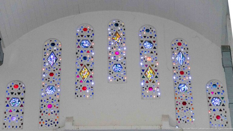 Seven narrow stained-glass windows of different heights arranged across a broad white interior wall.