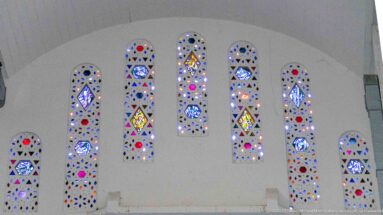 Seven narrow stained-glass windows of different heights arranged across a broad white interior wall.