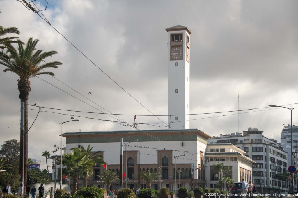 White civic building with a tall square clock tower, palms, overhead wires, and modern city blocks beneath dark clouds.