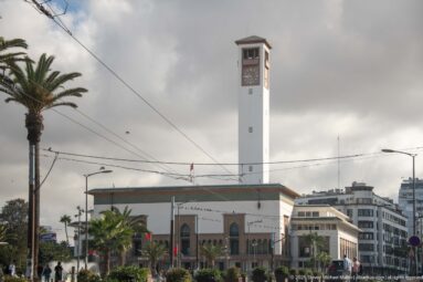White civic building with a tall square clock tower, palms, overhead wires, and modern city blocks beneath dark clouds.