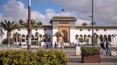 Large white Moroccan institutional building with arcades, a tall ochre gateway, green-tiled roof, flag, palms, and pedestrians.
