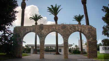 Freestanding rough-stone gateway with three round arches, tall palms, and a paved approach to low buildings.