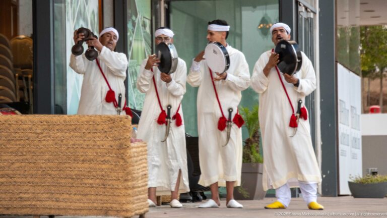 Four Moroccan musicians in white garments perform at an entrance with frame drums and metal percussion instruments.