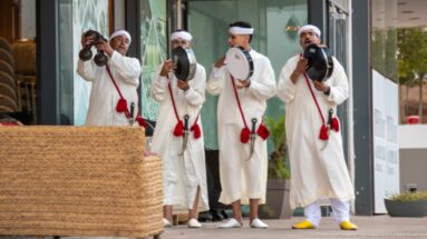 Four Moroccan musicians in white garments perform at an entrance with frame drums and metal percussion instruments.