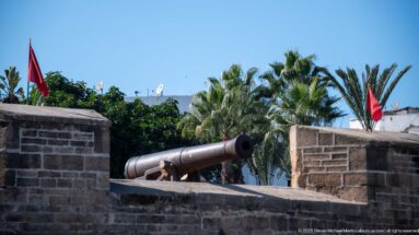 Dark iron cannon projects through a stone parapet with Moroccan flags and palm trees behind.