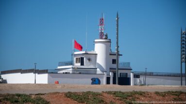 Low white lighthouse service building with Moroccan flag, antennas, communications mast, and rooftop equipment under a clear blue sky.