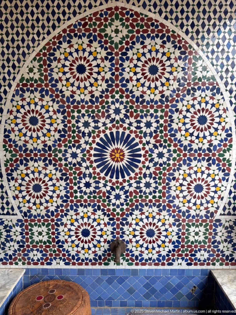 Large circular multicolored zellij mosaic with radiating rosettes and interlocking geometric patterns above blue tile.