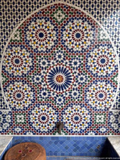 Large circular multicolored zellij mosaic with radiating rosettes and interlocking geometric patterns above blue tile.