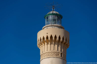 Close upward view of El Hank Lighthouse lantern with green trim, glass enclosure, projecting gallery, pointed corbels, and geometric ornament.
