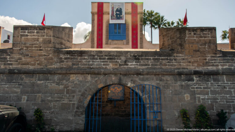 Stone wall with arched blue metal gate, Moroccan flags, and a bannered tower visible behind.