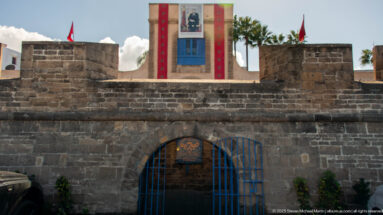Stone wall with arched blue metal gate, Moroccan flags, and a bannered tower visible behind.