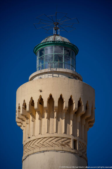 Tight view of El Hank Lighthouse lantern, balcony, pointed corbels, and patterned bands against a dark blue sky.