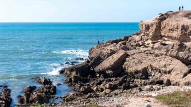 Rocky Atlantic shoreline with layered ledges, breaking waves, turquoise water, and small figures standing on the upper rocks.