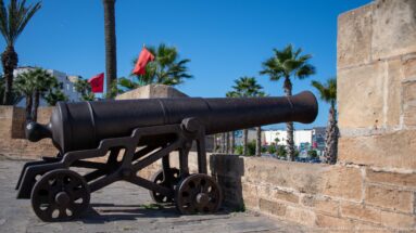Large iron cannon on a wheeled carriage beside stone walls, with Moroccan flags and palms behind.